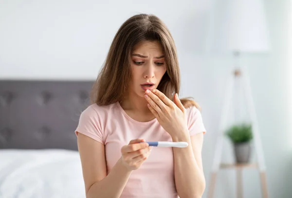 Mujer milenaria sorprendida comprobando la prueba de embarazo, sentada en la cama en casa, sorprendida con el resultado —  Fotos de Stock