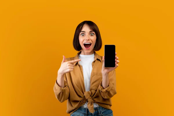 Портрет насолодженої жінки, що вказує на смартфон з порожнім екраном, позує на жовтому тлі, макет — стокове фото