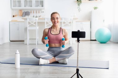 Neşeli Avrupalı bin yıllık sarışın fitness blogcusu webcam 'e bak ve dambıllarla antrenman videosu çek