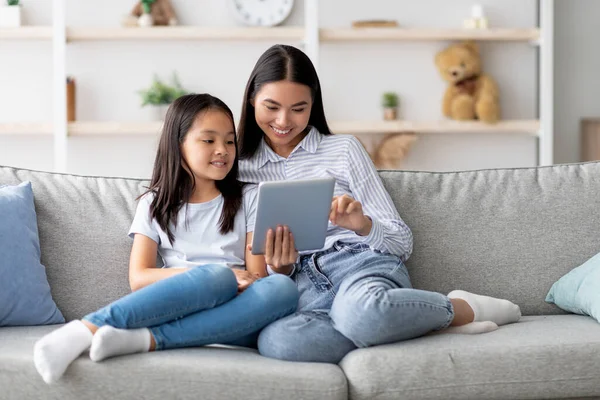 Happy young asian mom and daughter using digital tablet, watching videos or surfing internet, sitting on sofa — Stock Photo, Image