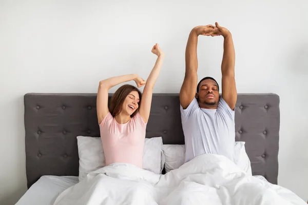 Multinacionális pár nyújtózkodik az ágyon ébredés után, otthon álmosnak érzik magukat — Stock Fotó