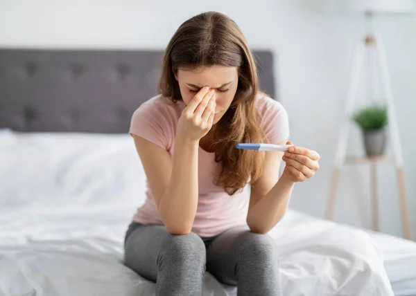 Sconvolta giovane donna che tiene il test di gravidanza, toccando il suo viso sotto stress sul letto a casa — Foto Stock