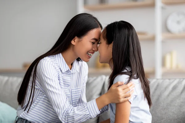 Söt asiatisk mor och dotter kramas och röra varandra med pannan sittande på soffan i vardagsrummet — Stockfoto