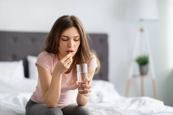 Ung kaukasisk dam som håller i glas vatten, tar piller på sängen, mår dåligt hemma, kopierar utrymme — Stockfoto