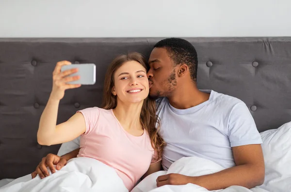 Evde yatakta selfie çekerken cep telefonuyla öpüşen genç bir çift. — Stok fotoğraf
