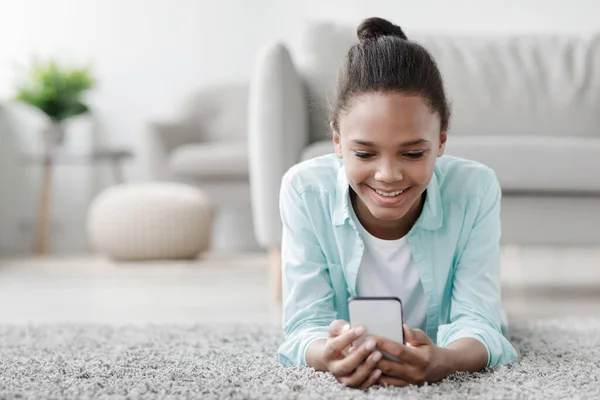 Felice ragazza afroamericana adolescente guarda il video online, chatta nei social network, si trova sul pavimento in soggiorno — Foto Stock