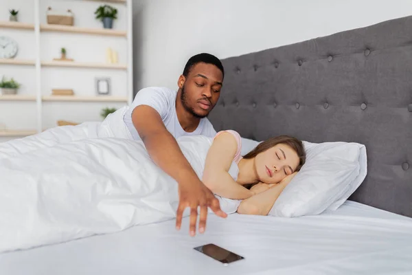 Hombre afro celoso revisando el teléfono móvil de sus novias dormidas en la cama en casa. Concepto de problemas matrimoniales — Foto de Stock