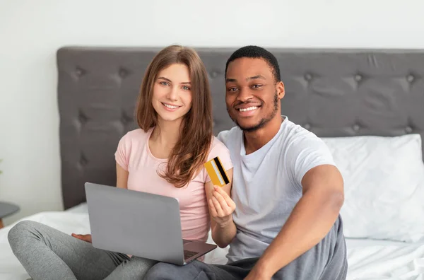 Attraktivt multinationellt par med kreditkort och laptop shopping online på sängen, gör kontaktlös betalning — Stockfoto