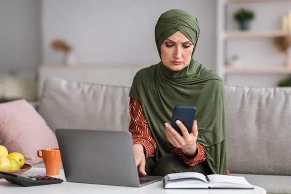 Schwere islamische Frau mittleren Alters benutzt Smartphone für Laptop im Haus — Stockfoto