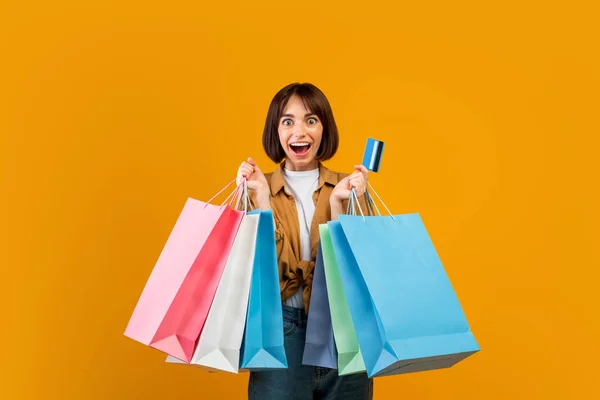 Koncepcja sprzedaży i rabatów. Podekscytowana kobieta z kartą kredytową i kolorowymi torbami na zakupy, żółte tło — Zdjęcie stockowe