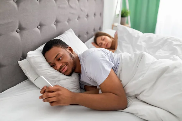 침대에 누워서 아내가 자고 있는 동안 핸드폰으로 애인에게 문자를 보내는 흑인 청년. 대인 관계의 불충실 — 스톡 사진