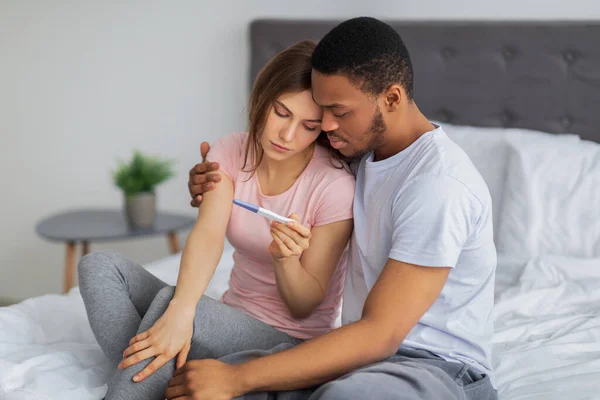 Unhappy black husband comforting his stressed young wife with negative pregnancy test on bed at home