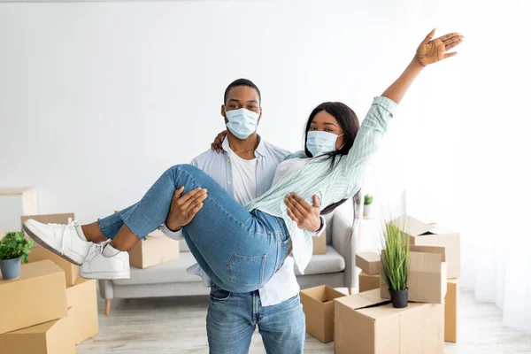 Celebrando el día de mudanza. Feliz pareja afroamericana en máscaras protectoras divirtiéndose, hombre sosteniendo a mujer en las manos — Foto de Stock