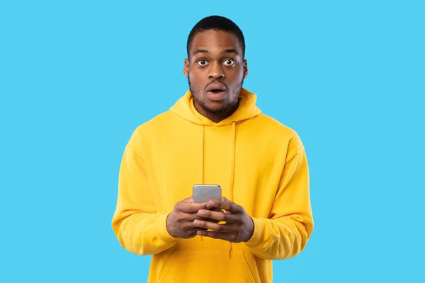 Impresionado chico afroamericano sosteniendo teléfono posando sobre fondo azul — Foto de Stock