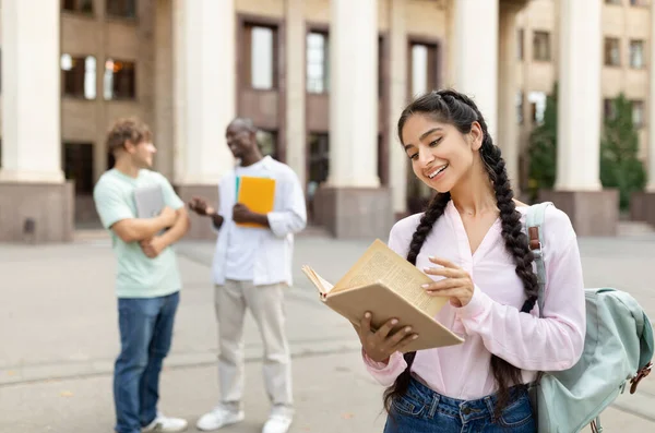 Feliz estudante universitário menina segurando livro, de pé ao ar livre no campus da universidade com seus colegas de classe em segundo plano — Fotografia de Stock