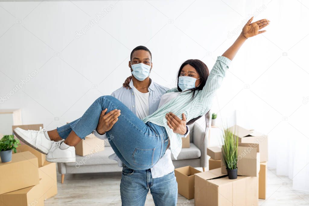 Celebrating moving day. Happy african american couple in protective masks having fun, man holding woman on hands