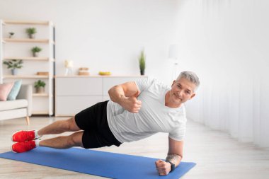 Güçlü, olgun bir adam evde çalışıyor, kalas yapıyor, baş parmağını kaldırıyor, oturma odasında kameraya gülümsüyor.