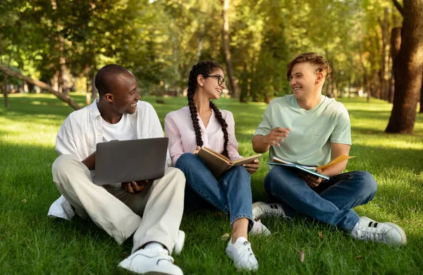 Счастливые иностранные студенты отдыхают в университетском городке, сидят на траве, разговаривают и используют ноутбук — стоковое фото