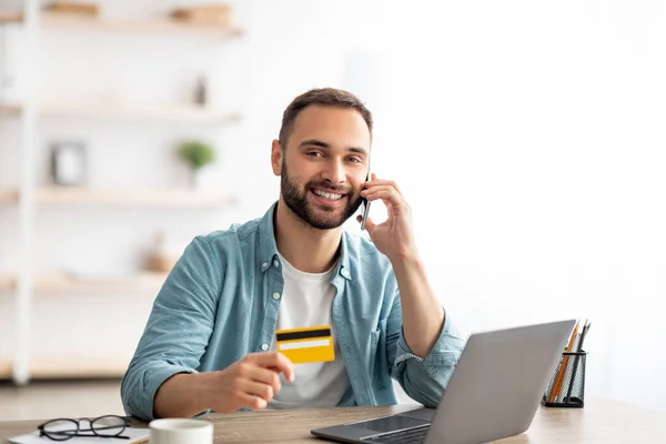 Χαρούμενος νεαρός αγοράζοντας πράγματα σε απευθείας σύνδεση, κάνοντας τηλεφώνημα, χρησιμοποιώντας φορητό υπολογιστή και πιστωτική κάρτα για ψώνια στο διαδίκτυο στο σπίτι — Φωτογραφία Αρχείου