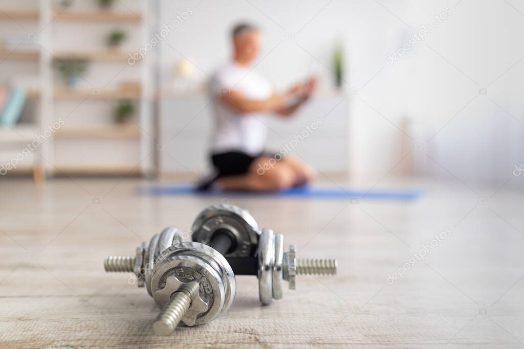 Home workout. Mature man training and using smartphone, sitting on mat at home, focus on dumbbells on foreground