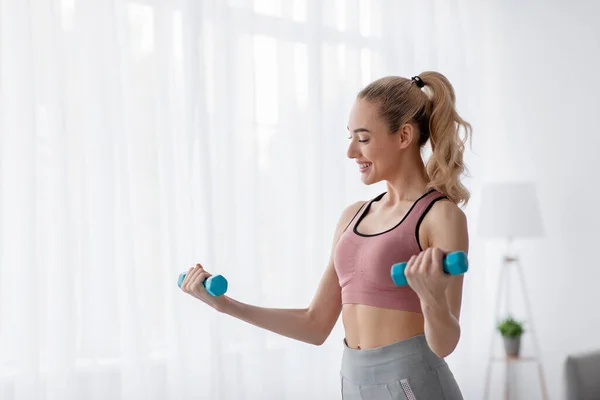Cheerful european cute young blonde woman lifts up dumbbells at home, work out alone, takes care of body — Stock Photo, Image