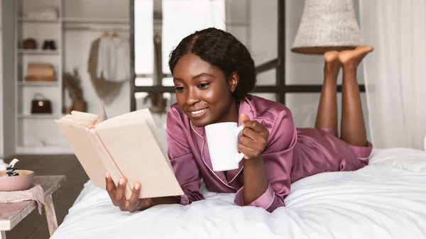 Μαύρη κυρία χαλαρωτικό βιβλίο ανάγνωσης πίνοντας καφέ που βρίσκεται στην κρεβατοκάμαρα — Φωτογραφία Αρχείου