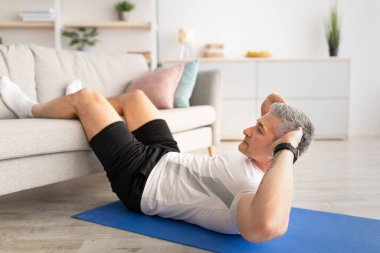 Canlılık için egzersiz. Atletik olgun bir adam karın kasları için egzersiz yapıyor, kanepeye ayak basıyor, evde paspas üzerinde egzersiz yapıyor.