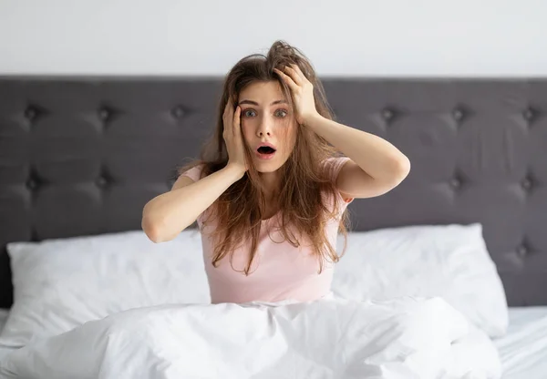 Sorprendida mujer milenaria sentada en la cama en casa, con la cabeza aterrorizada, llegando tarde al trabajo —  Fotos de Stock