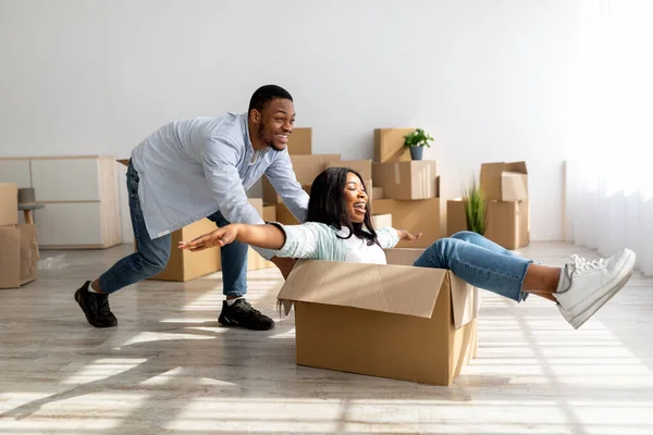 Casais negros alegres se divertindo ao se mudar para a nova casa, celebrando a realocação, andando em caixa de papelão — Fotografia de Stock