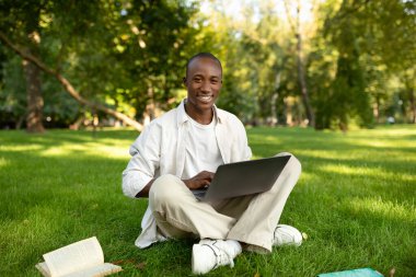 E-öğrenme. Açık havada dizüstü bilgisayar kullanan Afrikalı Amerikalı öğrenci, uzak online konferanslar veriyor, parkta çimlerin üzerinde oturuyor.