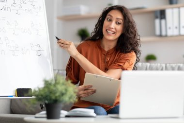 Dişi öğretmen elinde not panosu olan öğrencilere matematiği anlatıyor.