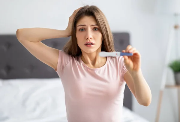 Scioccato giovane donna caucasica mostrando test di gravidanza negativi, tenendo la testa terrorizzata in camera da letto — Foto Stock