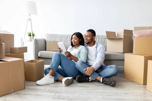 Αγαπώντας αφροαμερικανούς συζύγους χρησιμοποιώντας tablet, ψάχνοντας νέα έπιπλα, ενώ κάθεται ανάμεσα σε κουτιά από χαρτόνι, ελεύθερος χώρος — Φωτογραφία Αρχείου