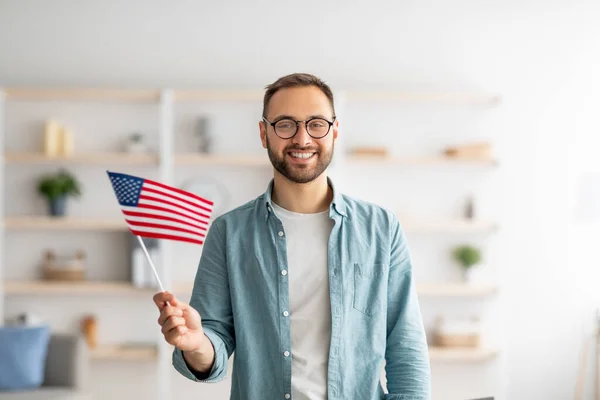 Snygg ung kille som håller amerikansk flagga och ler mot kameran inomhus. Utomlands utbildning och studentutbyte — Stockfoto