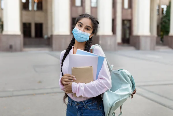 Estudiante india en máscara médica posando al aire libre con bloc de notas y libros, de pie cerca del edificio universitario —  Fotos de Stock