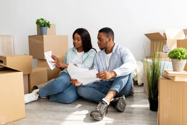Νεαρό μαύρο ζευγάρι σχεδιάζει ανακαίνιση στο σπίτι, τον έλεγχο σχέδια, ενώ κάθεται ανάμεσα σε κουτιά από χαρτόνι σε επίπεδη — Φωτογραφία Αρχείου