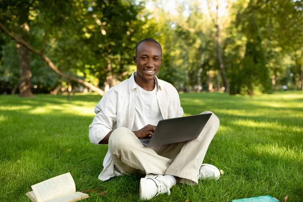 E-öğrenme. Açık havada dizüstü bilgisayar kullanan Afrikalı Amerikalı öğrenci, uzak online konferanslar veriyor, parkta çimlerin üzerinde oturuyor.