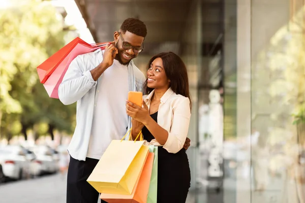 Portret van gelukkig zwart paar met behulp van telefoon met boodschappentassen — Stockfoto