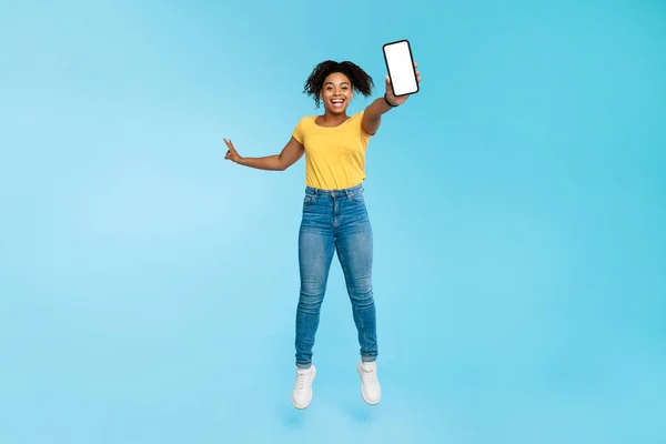 Joven dama negra llena de alegría saltando con un teléfono inteligente vacío sobre fondo de estudio azul, maqueta — Foto de Stock