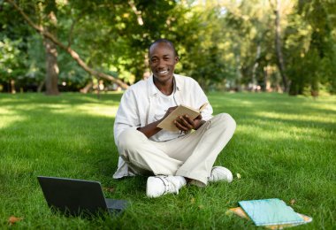 Mutlu Afro-Amerikalı çocuk parkta dizüstü bilgisayarla ders çalışıyor, kampüste oturuyor ve notlar alıyor.