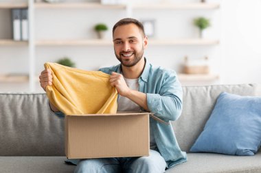 Karton paketleri açan mutlu beyaz adam, başarılı alışverişlerden memnun, evde yeni bir kazak çıkarıyor.