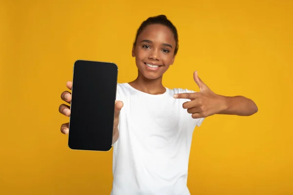 흰색 티셔츠를 입은 쾌활 한 미국 여학생 이 백지 스크린을 가진 스마트폰을 손가락으로 가리키며 말한다. — 스톡 사진