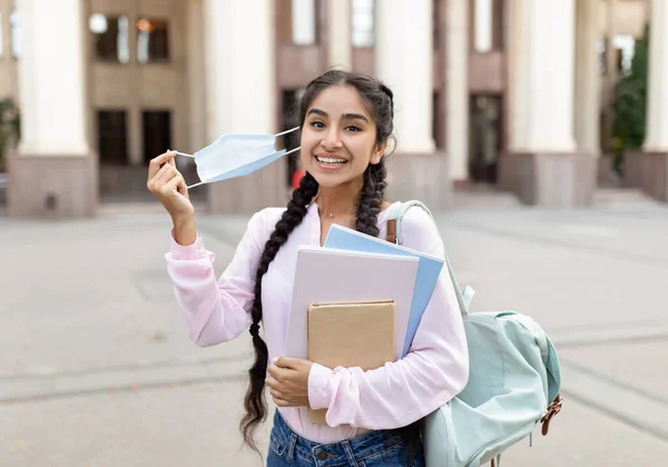 Estudante indiano feliz removendo máscara médica, posando ao ar livre com blocos de notas perto do prédio da universidade — Fotografia de Stock