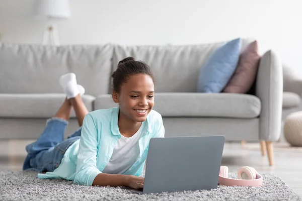 快乐的年轻黑人女生带着笔记本电脑在家里学习，与朋友聊天，看视频，躺在地板上 — 图库照片