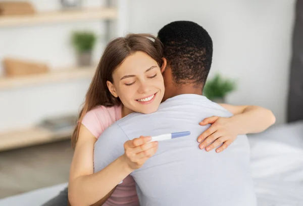 Fröhliche junge Frau umarmt ihren schwarzen Mann, hält positiven Schwangerschaftstest zu Hause — Stockfoto