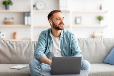 Kulaklıklı gülümseyen genç adam evdeki internet iletişimi için laptop kullanıyor.