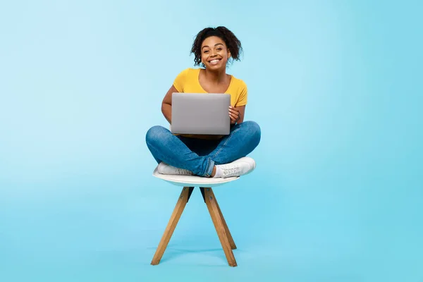 Joven mujer negra alegre que trabaja en línea, sentado en la silla y usando el ordenador portátil en fondo azul del estudio, longitud completa — Foto de Stock