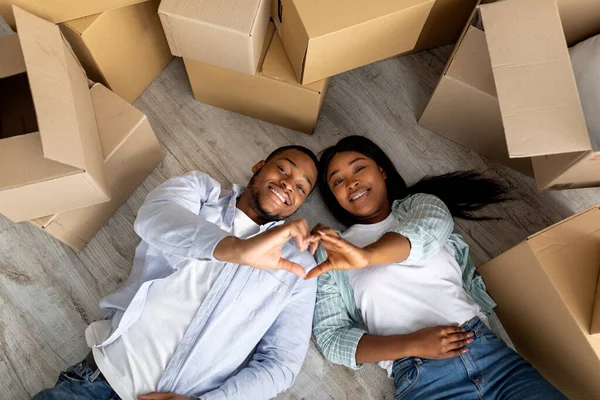 Власність будинку. Веселі чорні подружжя роблять жест форми серця, лежачи на підлозі біля коробки в день переселення — стокове фото