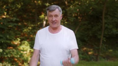 Aktif kıdemli bir adam dışarıda halterlerle egzersiz yapıyor, pazılarını parkta çalıştırıyor, çekim takibini yapıyor.