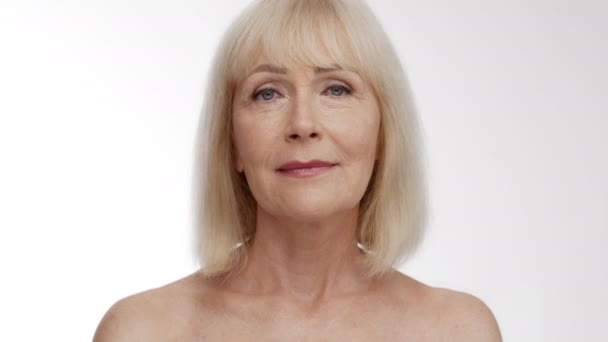 Beauty portrait of gorgeous senior blonde lady with bare shoulders smiling to camera, white studio background — Stock Video